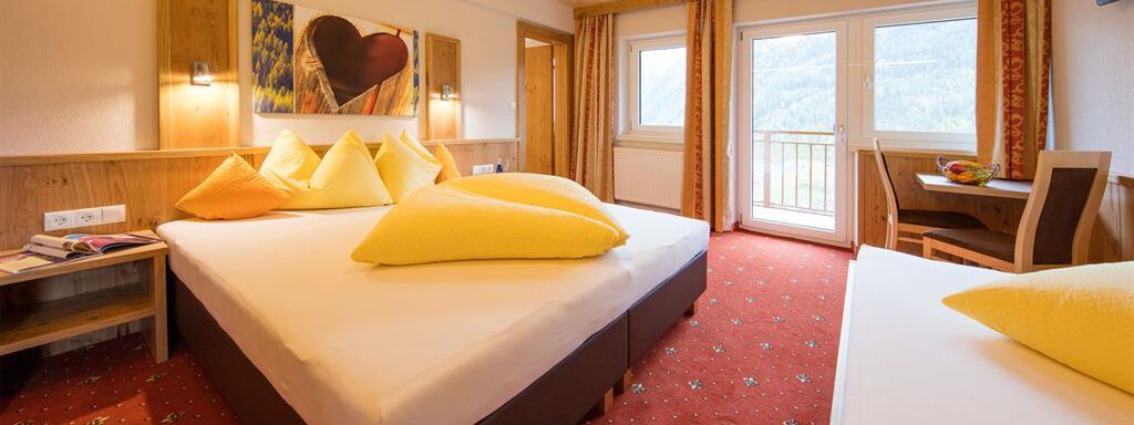 A cozy hotel room with two beds and yellow pillows. Large windows offer a view outside and plenty of daylight. - Haus Stefanie - Sölden