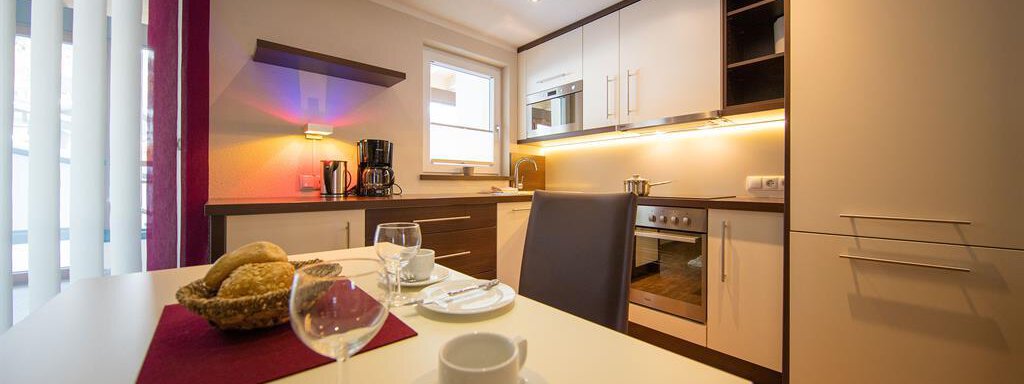 A modern kitchen with bright furniture and warm light. The table is beautifully set with dishes and a fruit basket. - Haus Stefanie - Sölden