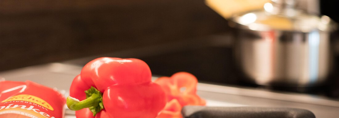 A red bell pepper lies on a cutting board next to a knife. In the background, a pot is visible on the stove. - Haus Stefanie - Sölden