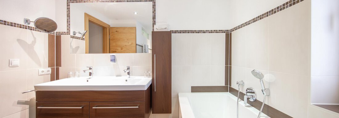 A modern bathroom with a double sink, wooden cabinet, and a bathtub. The walls are light, and the tiles have a simple design. - Haus Stefanie - Sölden