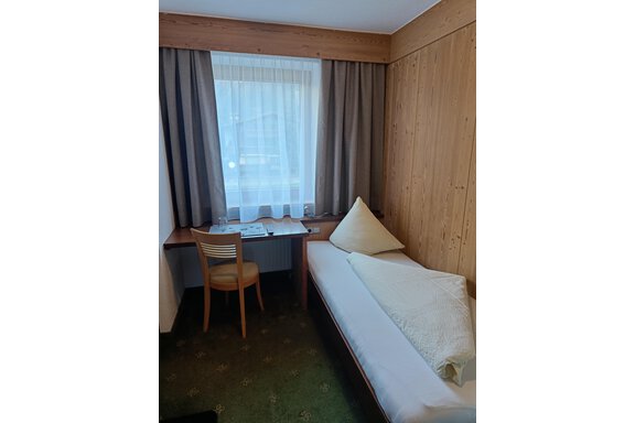 A small room with wooden paneling and a window that lets in a lot of light. There is a bed and a table with a chair. - Haus Tanja - Sölden