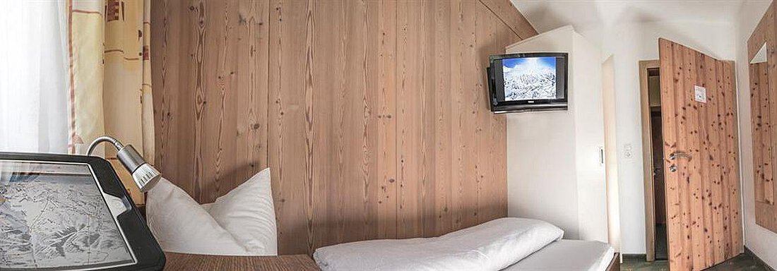 Einzelzimmer A simple room with wooden paneling and a single bed. A television is mounted on the wall, and there is a view of a door. - Haus Tanja - Sölden