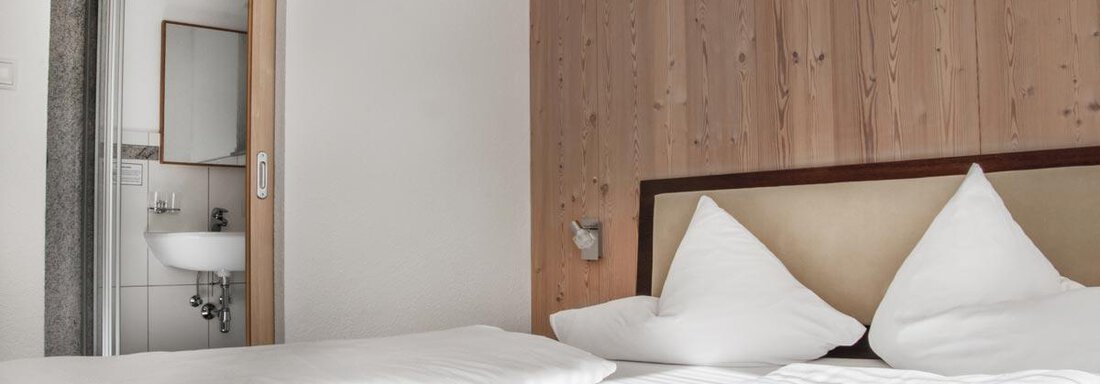 A bright bedroom with a double bed and pillows. In the background, a bathroom area is visible. - Haus Tanja - Sölden