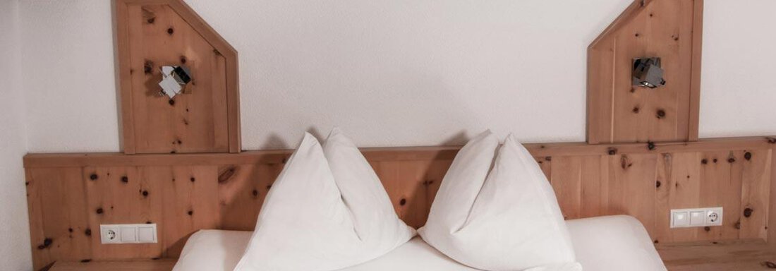 A cozy bedroom with a double bed and high pillows. The walls are made of light wood with decorative elements. - Haus Tanja - Sölden