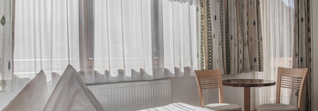 A bright room with large windows and curtains. There is a bed and a small table with chairs. - Haus Tanja - Sölden