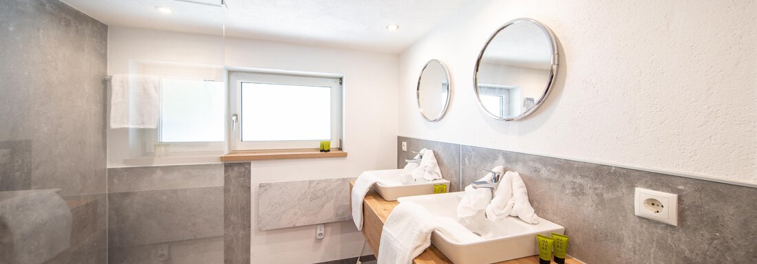 A modern bathroom with two sinks and large mirrors. The walls are in neutral tones and there is a shower in the corner. - Haus Valtelas - Sölden