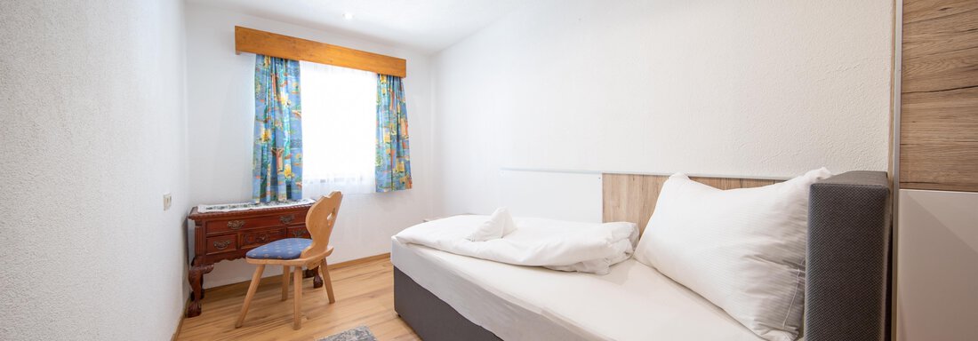 A simple room with a single bed, a table, and colorful curtains. The room has bright walls and a wooden floor. - Haus Valtelas - Sölden