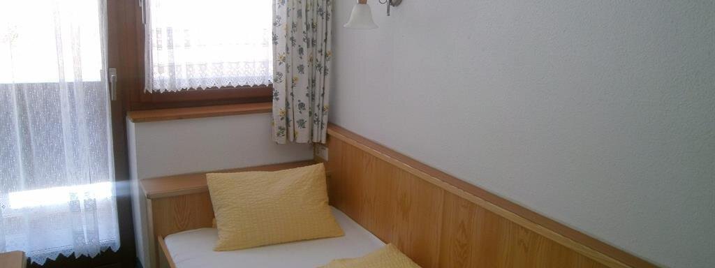 A simple room with a single bed and a yellow blanket. Light shines through the windows, and a cozy atmosphere is present. - Hirschenhof - Sölden