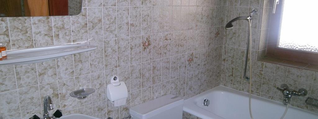 A bathroom with a bathtub, a sink, and a toilet. The walls are tiled and there is a window for daylight. - Hirschenhof - Sölden