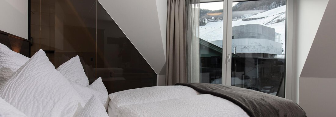 A modern bedroom with a comfortable bed and decorative pillows. The view from the window shows snow-capped mountains. - Home4You - Sölden