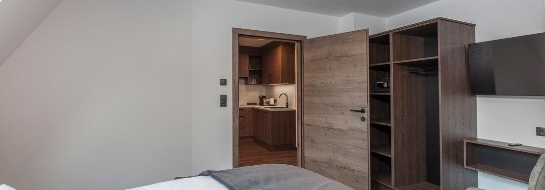 A modern hotel room with a comfortable bed and a bright, friendly atmosphere. In the background, a refrigerator and a kitchenette are visible. - Home4You - Sölden