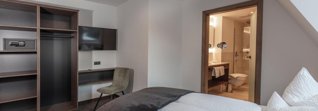 A modern hotel room with a large bed, a desk, and a television. In the background, there is an adjoining bathroom. - Home4You - Sölden