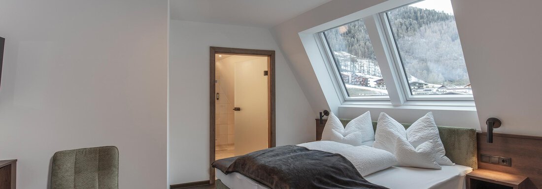 A modern bedroom with a large bed and white pillows. The windows offer a view of the surrounding mountains and the shower is also visible. - Home4You - Sölden