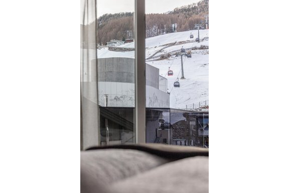A view out the window of a snowy landscape with ski lifts. In the background, mountains and a modern building can be seen. - Home4You - Sölden