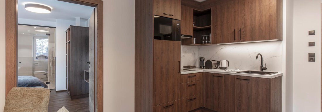 A modern kitchen with dark wooden furniture and white work surfaces. In the background, a bright living area is visible. - Home4You - Sölden