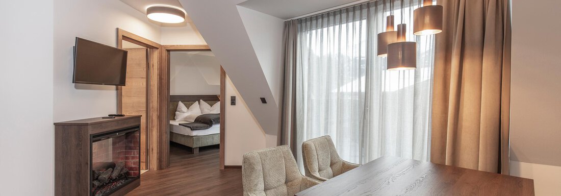 A modern living and bedroom with a table, two chairs, and a television on the wall. The large windows let in plenty of light and create a cozy atmosphere. - Home4You - Sölden