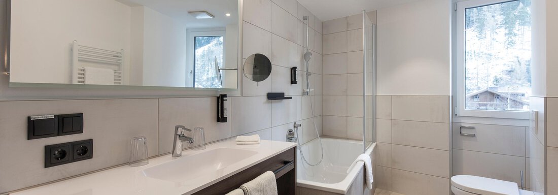 A modern bathroom with a bathtub and a sink. The walls are brightly tiled and there is a window overlooking the outside. - Home4You - Sölden