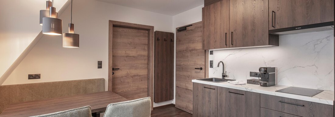 A modern kitchen with wooden cabinets and a light countertop. The dining area includes a table with two chairs and stylish pendant lights. - Home4You - Sölden