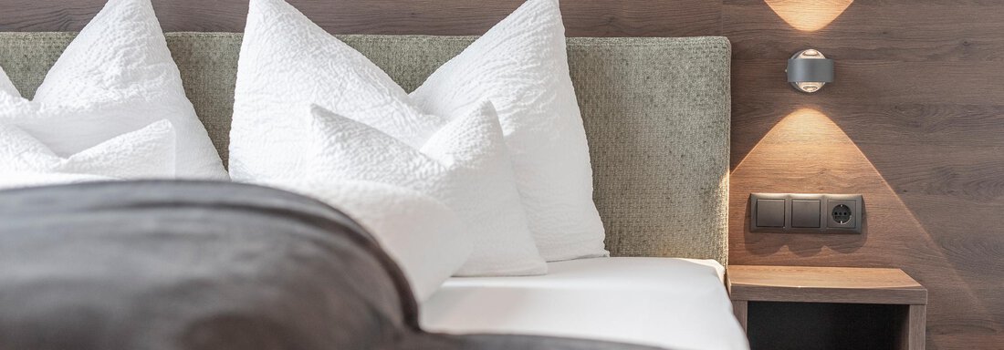 A modern bedroom with a cozy bed and many white pillows. Stylish lamps are on the wall and there is a nightstand with power outlets. - Home4You - Sölden