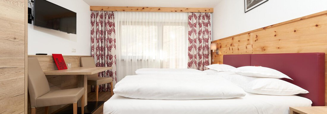 A modern hotel room with a double bed, a desk, and a television. Large windows let in plenty of light and create an inviting atmosphere. - Hotel am Hof - Sölden