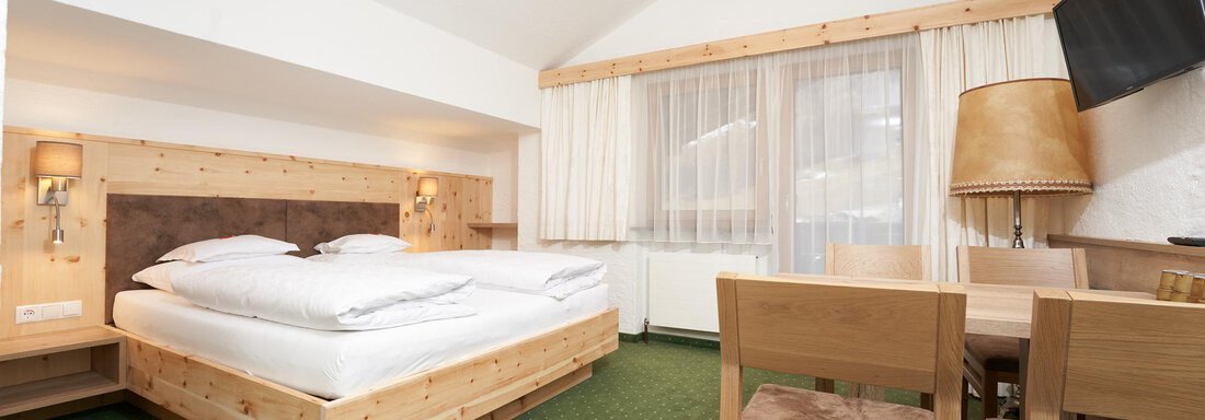 A modern hotel room with a double bed and a small dining area. The furniture is made of light wood and the floor is covered with green carpet. - Hotel am Hof - Sölden