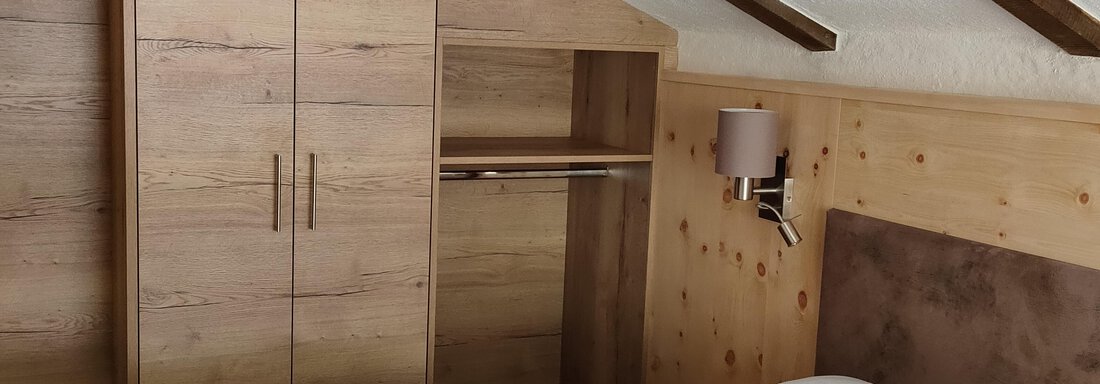 A cozy bedroom with wooden paneling and sloped ceilings. The room is furnished with a bed, a wardrobe, and a bedside lamp. - Hotel am Hof - Sölden