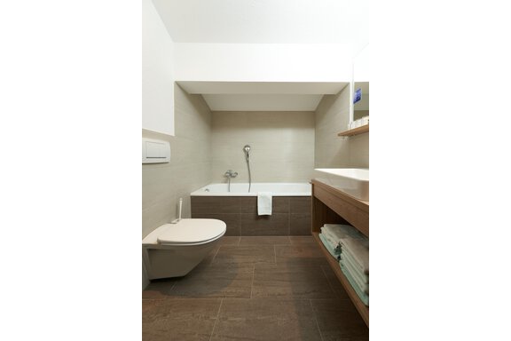 A modern bathroom with a bathtub and a toilet. The walls are painted in neutral tones, and there is a wooden shelf with fresh towels. - Hotel am Hof - Sölden