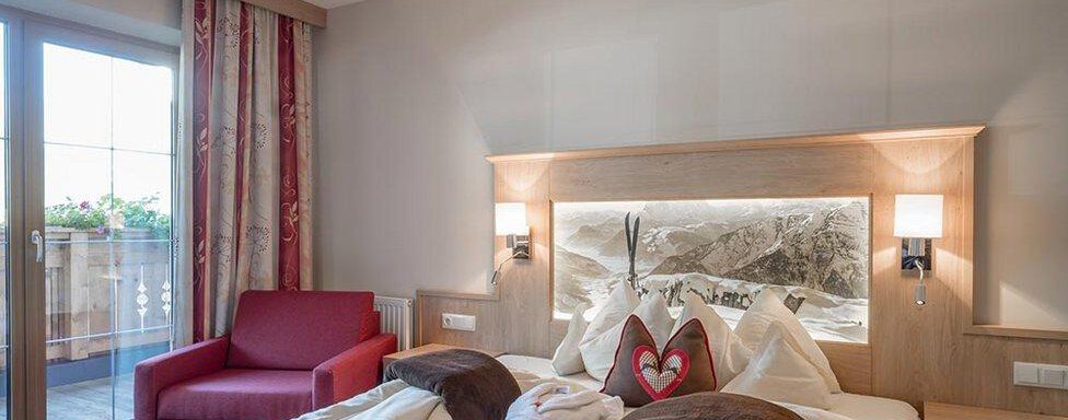 Schlafzimmer Hotelappartement | © Hotel Elisabeth (CC C) A cozy bedroom with a large bed and elegant wooden furniture. A red armchair and a balcony with windows create a bright atmosphere. - Hotel Elisabeth - Sölden