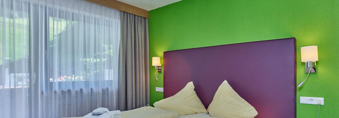 Doppelzimmer Sölden A modern bedroom with a large double bed and colorful walls. Plenty of light from the windows and fresh amenities are visible. - Hotel Erhart - Sölden