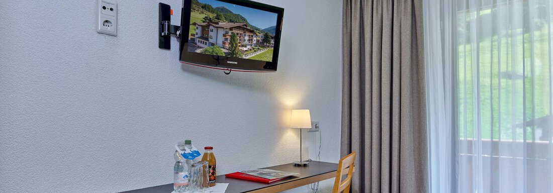 A cozy hotel room area with a desk, a television, and a chair. A window with curtains lets in natural light. - Hotel Erhart - Sölden