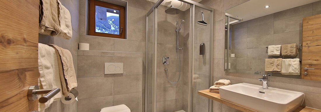 A modern bathroom with a shower and a large sink. The walls are painted in neutral shades of gray, and there are wooden details for a warm accent. - Haus Söldenblick - Sölden