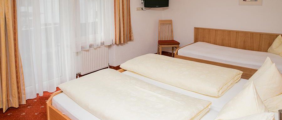 Schlafzimmer A simple hotel room with two beds and plenty of pillows. There is a window with curtains and a small television on the wall. - Hotel Garni Ehrenreich - Sölden