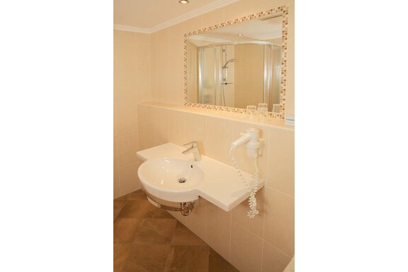 A modern bathroom with a white sink and a large mirror. The walls are tiled in light colors, and there is a shower in the background. - Hotel Garni Ehrenreich - Sölden