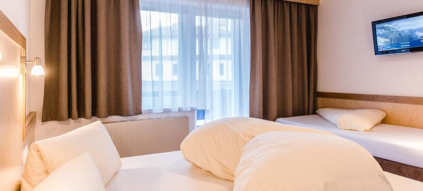 A bright bedroom with two beds and large windows. The decor is simple and modern, with curtains and a television on the wall. - Hotel Garni Ehrenreich - Sölden