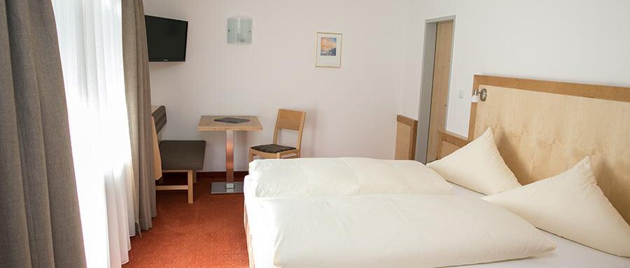 Doppelzimmer A bright room with a double bed and large windows. There is a table with chairs and a television on the wall. - Hotel Garni Ehrenreich - Sölden