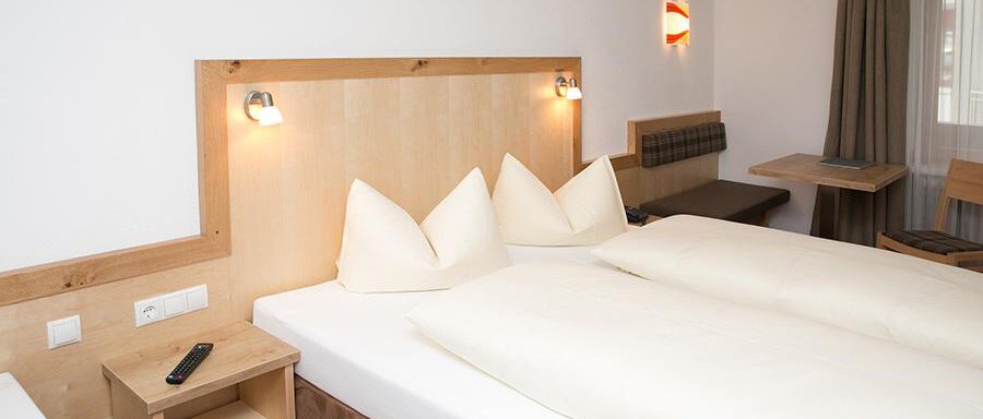 A modern hotel room with a double bed and two pillows. There is a simple wood and fabric decor, as well as a small table with chairs. - Hotel Garni Ehrenreich - Sölden