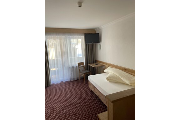 Einzelzimmer A simple bedroom with a bed, a table, and a chair. There is a television and large windows with curtains. - Hotel Garni Ehrenreich - Sölden