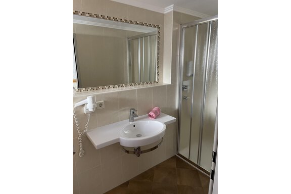A modern bathroom with a sink, mirror, and a shower. The walls are light-tiled and the floor is brown. - Hotel Garni Ehrenreich - Sölden