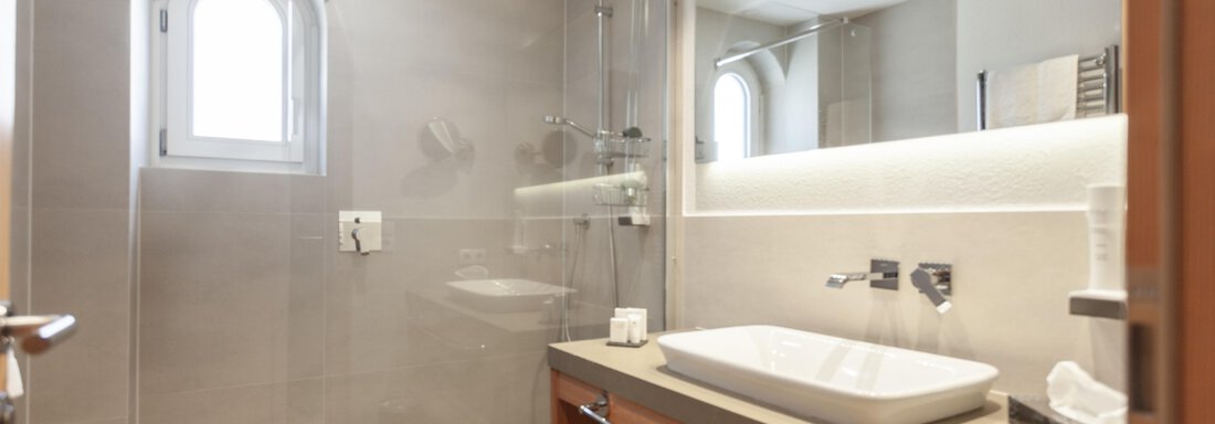 A modern bathroom with a walk-in shower and an elegant sink. The walls are kept in neutral tones and there is plenty of light through the window. - Hotel Hubertus - Sölden
