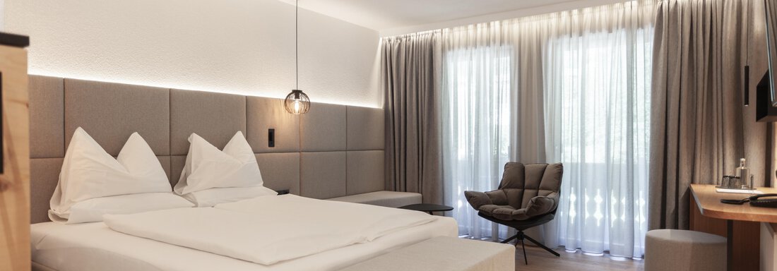 A modern bedroom with a large bed, elegant furnishings, and windows with curtains. The room has a relaxed atmosphere and plenty of light. - Hotel Hubertus - Sölden