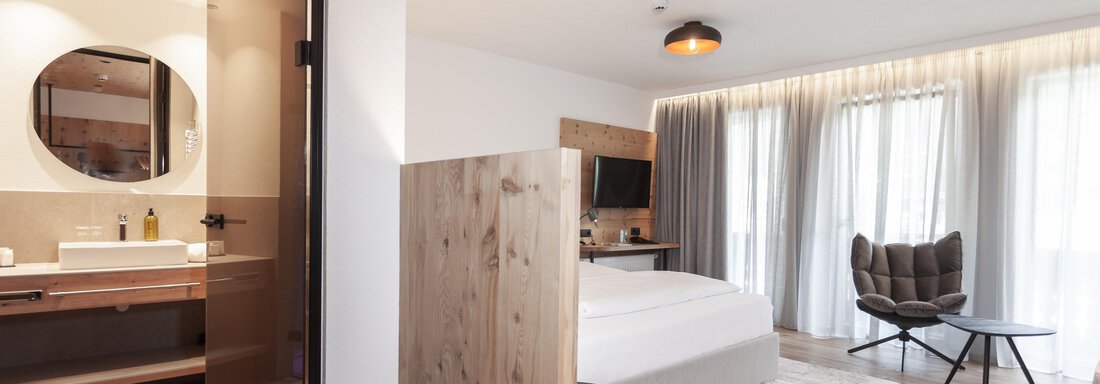 A modern bedroom with a cozy bed and a comfortable armchair. Large windows provide plenty of daylight. - Hotel Hubertus - Sölden
