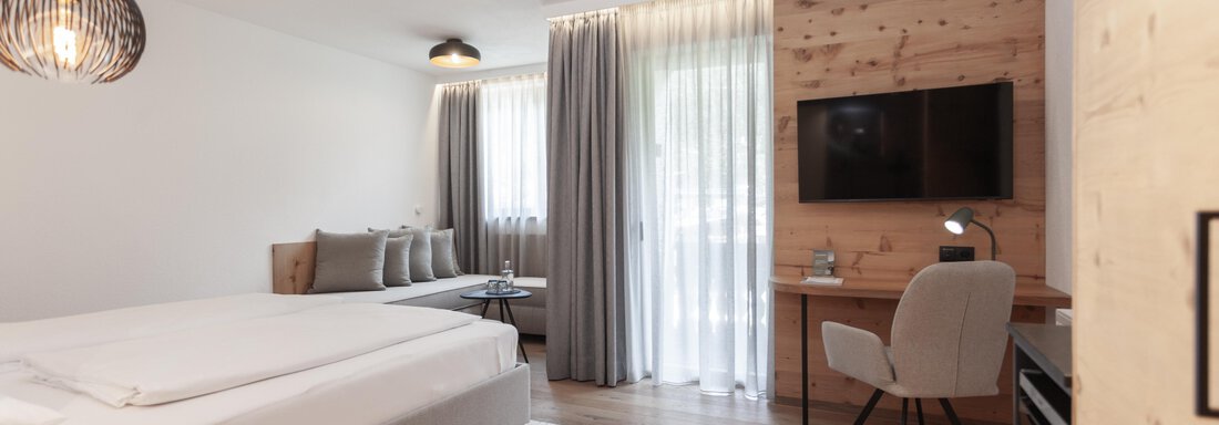 A modern and cozy hotel room with a double bed and wood paneling. There is a desk, a television, and large windows that let in plenty of light. - Hotel Hubertus - Sölden