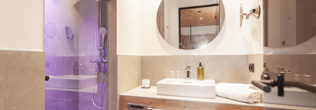 A modern bathroom with a shower and a large mirror. There is a wooden washbasin with toiletries and a towel. - Hotel Hubertus - Sölden