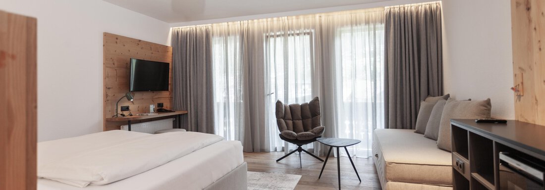 A modern hotel room with a large bed and a comfortable seating area. Large windows provide plenty of daylight and an inviting atmosphere. - Hotel Hubertus - Sölden