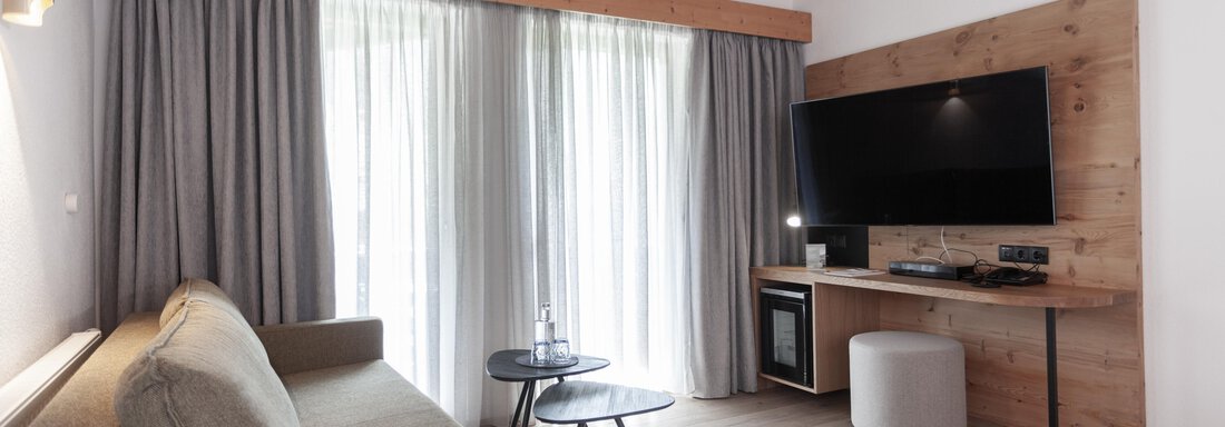 A modern living room with a comfortable sofa and a flat-screen television.  
Large windows with curtains let in plenty of light. - Hotel Hubertus - Sölden
