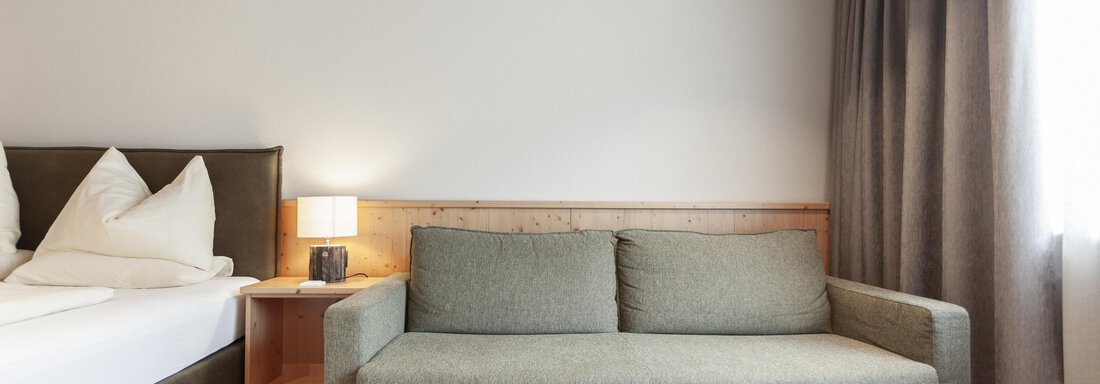 A modern room with a cozy sofa and a large bed. The walls are light and there are curtains that gently filter the light. - Hotel Hubertus - Sölden