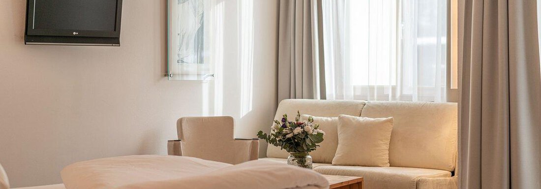 A bright room with a cozy sofa and a small table. In the background, there is a television and large windows with curtains. - Hotel Liebe Sonne - Sölden