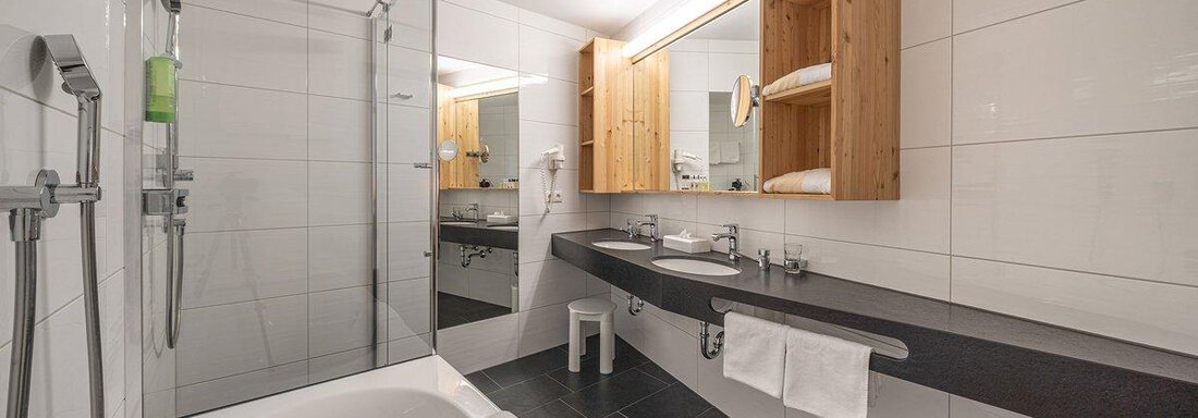 A modern bathroom with a shower and a bathtub. The walls are light tiled, and there are wooden shelves for towels. - Hotel Liebe Sonne - Sölden