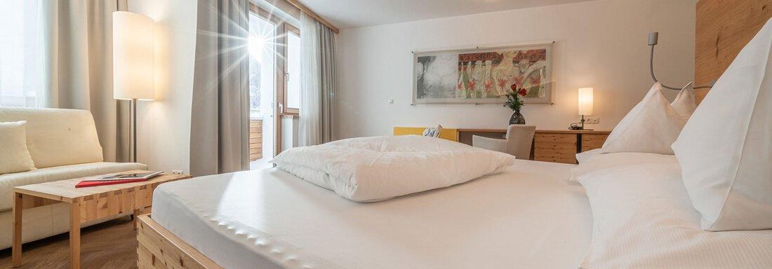 A cozy hotel room with a large bed and light wood interior. The windows let in plenty of light, and there is a small living area with a sofa. - Hotel Liebe Sonne - Sölden