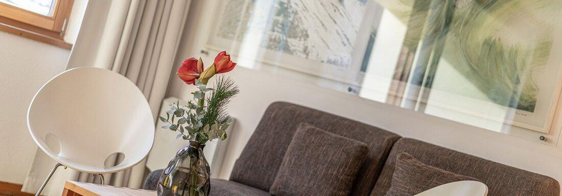 A modern living room with a gray sofa and a wooden table. On the table, there is a vase of red flowers. - Hotel Liebe Sonne - Sölden